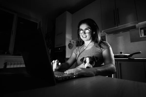 Une femme souriante avec des lunettes est assise dans le noir à un ordinateur sans fil dans la cuisine avec un chiot de Jack Russell Terrier sur les genoux. Fille avec son petit chien regarder un film sur un ordinateur portable à la maison . — Photo