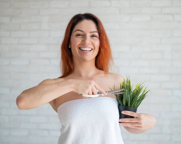 A smiling red-haired woman in a white terry towel cuts a green plant in a pot with scissors. Imitation of armpit depilation. Hair removal concept. Procedure for hair removal of unwanted vegetation.