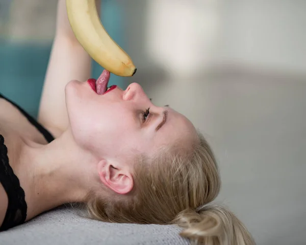 Blondinen i svarta spetsunderkläder äter erotiskt en banan. Attraktiv kvinna med sensuella röda läppar suger sexuellt och slickar en banan. Oral njutning. — Stockfoto