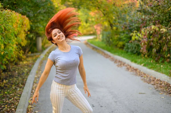 一个红头发的漂亮女人正在街上走着跳舞。穿着灰色T恤和条纹腿的快乐女孩散步，在秋天的公园里玩得很开心。照片在动. — 图库照片