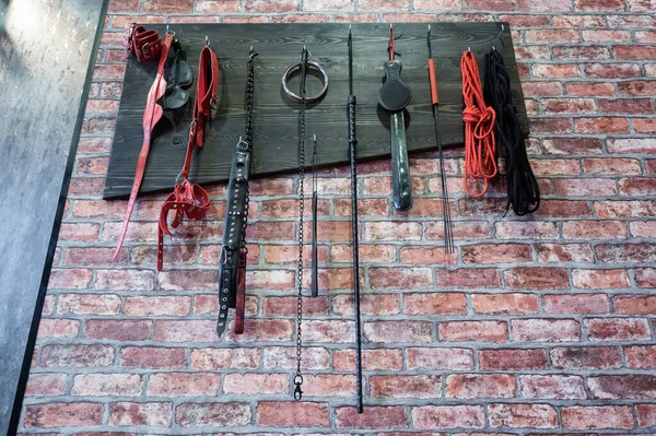 Ett set med olika läderfransar för sexuella njutningar hänger på en trähängare på en tegelvägg. Sexutrustning i rött och svart för rollspel. BDSM-rummet. — Stockfoto