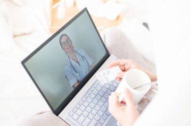 Bir kadın yatağa uzanır ve boğaz ağrısı için sprey tutar. Dost canlısı bir doktor hasta bir hastayla video bağlantısıyla konuşur. Laptop ekranında terapist. Karantina sırasında çevrimiçi tıbbi konsültasyon.
