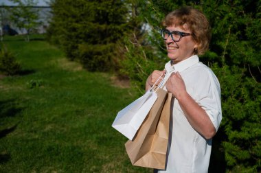 Gözlüklü mutlu yaşlı kadın parkta yürürken çevre dostu bir paket tutuyor. Keyifli yaşlı bir kadın, açık havada kağıt torbalarla alışveriş yapıyor..