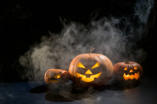 Tři strašidelné halloween dýně s vyřezávaným zářivým úšklebkem na černém pozadí. Ručně vyrobená hlava z lucerny se svíčkou ve tmě mezi mlhou. Trick or treat. Pohlednice. — Stock fotografie