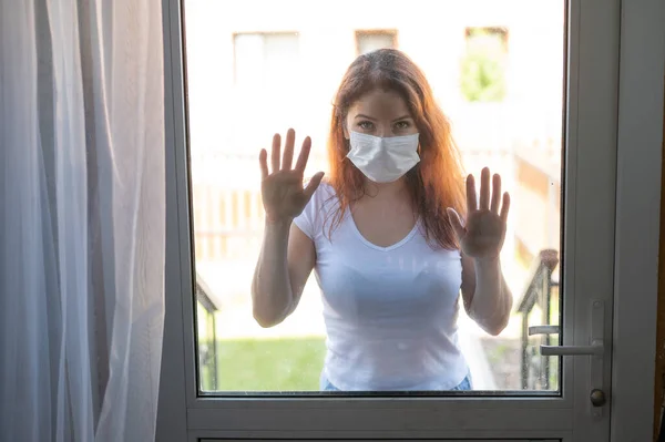 一个戴着医疗面罩的红头发女人靠着前玻璃门。检疫期间,一个女孩站在窗外.科罗纳威斯流行病社会距离. — 图库照片