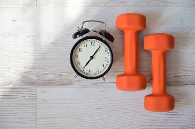 Bir çift turuncu dambıl ve çalar saatin üst görüntüsü. Sabahın köründe spor yapma fikri. Evde kendini eğitme zamanı..