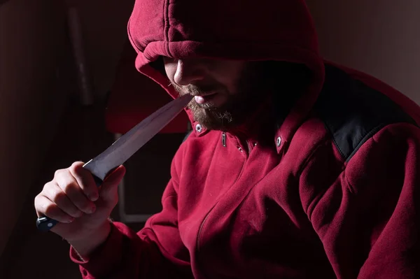 一个戴着红色头巾的危险的大胡子男人在他的脸旁边拿着一把锋利的小刀。黑暗中拿着刀的罪犯或杀人犯. — 图库照片