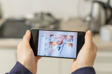 Ciddi sakallı bir doktor bir hastayla akıllı telefondan görüntülü arama yoluyla iletişim kurar. Çevrimiçi tıp konsepti. Mutfaktaki kadın cep telefonundan terapiste gidiyor..