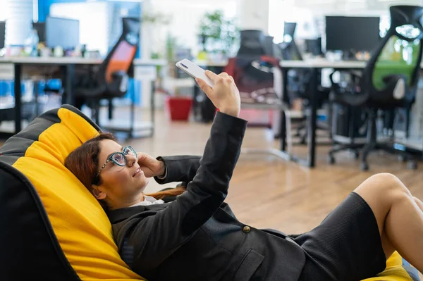 Una mujer de negocios en un traje está descansando mientras está sentada en una cómoda bolsa de silla y tomando una selfie. Una empleada utiliza un teléfono inteligente y se relaja mientras está acostada en una silla de frijol durante un descanso en el trabajo . —  Fotos de Stock