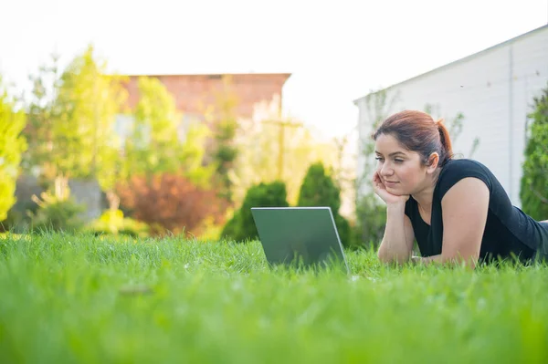 Uma mulher se deita de barriga no gramado da casa de campo e usa em um laptop. Uma freelancer focada trabalha remotamente ao ar livre. A menina está estudando no computador . — Fotografia de Stock