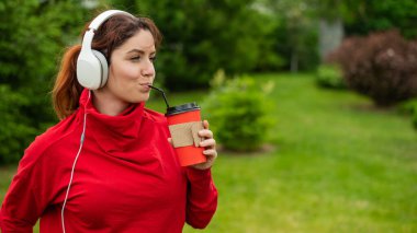 Kızıl saçlı kadın kulaklıkla müzik dinliyor ve parkta pipetle kahve içiyor. Bir kız güneşli bir yaz gününde yürüyüş yapmaktan zevk alır ve bir paket kahve alır. Akıllı telefona bağlı kulaklıklar..