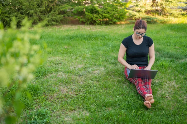 Uma mulher ruiva senta-se na grama verde em um parque e está comprando on-line. A menina mantém uma distância social e trabalha ao ar livre em um laptop. Um estudante está estudando remotamente em um computador . — Fotografia de Stock