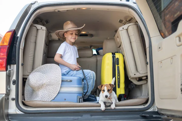 Glücklicher Junge mit Cowboyhut und Welpe Jack Russell Terrier reisen mit dem Auto. Ein Kind und ein lustiger kleiner Hund sitzen im Kofferraum und sind bereit für die Sommerferien. Unabhängiges Reisen. Beste Freunde. — Stockfoto