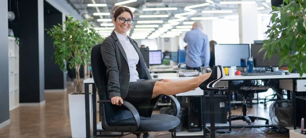 Feliz mujer de negocios haciendo ejercicios musculares abdominales en una oficina de espacio abierto. Una empleada pelirroja sonriente con una falda y tacones altos está muriendo de una silla en el lugar de trabajo. Fitness en el trabajo . —  Fotos de Stock