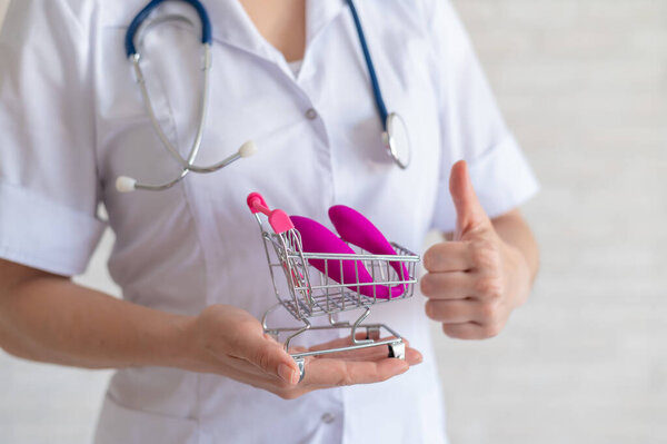 The faceless gynecologist recommends buying a clitoral vaginal vibrator to maintain womens health. The doctor holds a mini trolley and a masturbator for vivid orgasms. Gesture thumb.