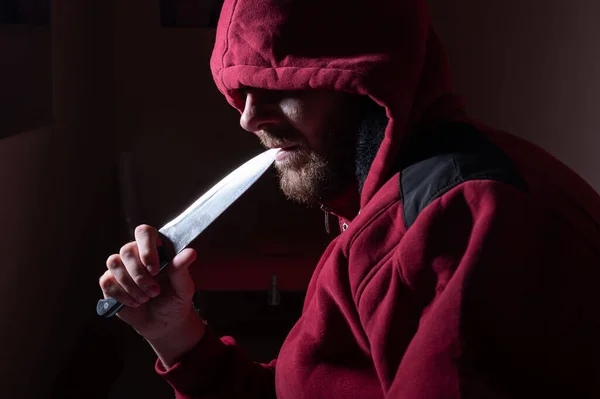 빨간 후드를 쓰고 있는 위험 한 수염을 가진 한 남자가 날카로운 칼을 얼굴 가까이에 들고 있다. 어둠 속에서 칼을 든 범죄자나 살인자. — 스톡 사진