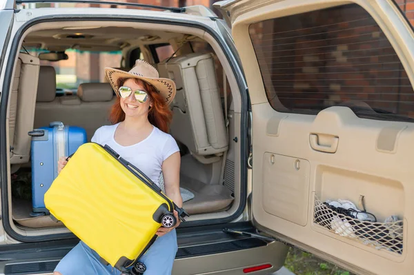 Glückliche rothaarige Frau mit Cowboyhut und Sonnenbrille auf einer Autofahrt durch das Land. Mädchen im Kofferraum mit gelbem Koffer, bereit für den Sommerurlaub. Unabhängiges Reisen. — Stockfoto