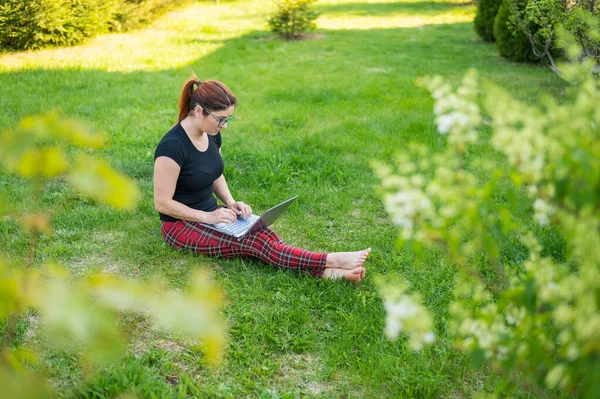 Uma mulher ruiva senta-se na grama verde em um parque e está comprando on-line. A menina mantém uma distância social e trabalha ao ar livre em um laptop. Um estudante está estudando remotamente em um computador . — Fotografia de Stock