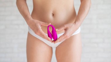 A faceless woman in white panties holds a curved pink clitoral vaginal vibrator. Female sex toy for masturbation. Self-gratification. Demonstrates dildo.
