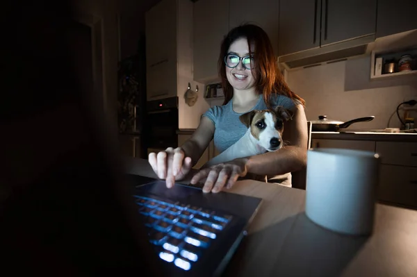 Une femme souriante à lunettes est assise à un ordinateur sans fil dans la cuisine avec un chiot de Jack Russell Terrier à genoux. Fille pigiste travaille à un ordinateur portable à la maison et boit du café . — Photo