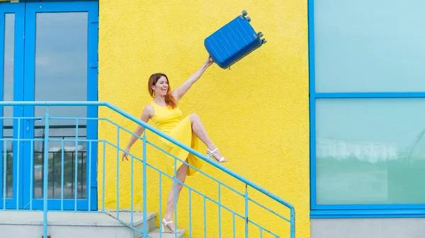 Счастливая рыжая женщина в жёлтом платье спускается по лестнице и радостно размахивает голубым чемоданом. Девушка готовится к путешествию. Летние каникулы. — стоковое фото