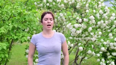 Güzel kızıl saçlı kadın hapşırır ve parkta çiçek açan bir elma ağacının yanında dururken burnunu ovuşturur. Mevsimsel alerjik rhinitis. Kızın polene alerjisi var..
