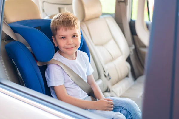 Little Caucasian boy sits in a blue child safety seat. Traveling with a child in a car with a leather interior. Portrait of a cute preschooler wearing a seat belt and ready to ride. — Stock Photo, Image