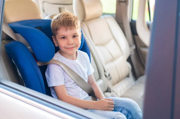 El pequeño chico caucásico se sienta en un asiento azul de seguridad para niños. Viajar con un niño en un coche con un interior de cuero. Retrato de un niño en edad preescolar lindo con un cinturón de seguridad y listo para montar . —  Fotos de Stock