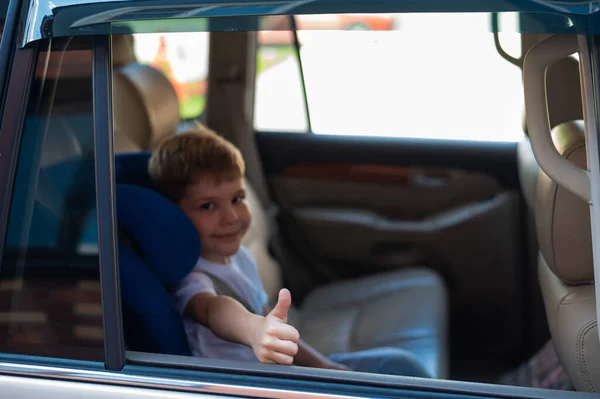Menino caucasiano feliz sentado em um assento de segurança infantil azul e mostrando o polegar. Viajar com uma criança em um carro com um interior de couro. Retrato de um pré-escolar bonito usando um cinto de segurança e pronto para montar — Fotografia de Stock