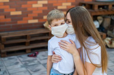 Kadın açık havada maske takmış oğluna sarılıyor. Annesiyle küçük mutlu çocuk tecrit sırasında arka bahçede yürüyor. Koronavirüsün yayılmasına karşı koruma. Grip salgını.