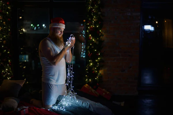 Komický obrázek Santa Clause ve spodním prádle na Štědrý den. Muž v obleku Santa Clause drží na Nový rok věnec se světly. Parodie na ženské focení. — Stock fotografie