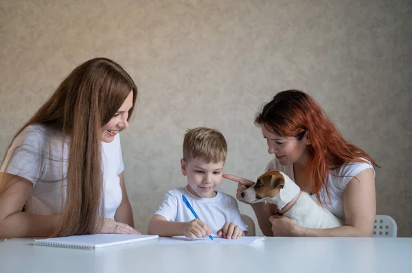 Happy family stays at home. Two women help the boy do school homework. Lesbian couple sitting at the table with their son and a cheerful puppy. Same-sex marriage with a child. Distance learning.