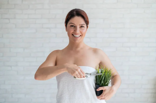 Vackra leende kvinna i en vit frottéhandduk skär en grön växt i en kruka med sax. Imitation av armhålan avlivning. Koncept för hårborttagning. Förfarande för hårborttagning av oönskad vegetation. — Stockfoto