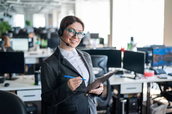 A friendly support woman with glasses and a business suit is talking on a headset with a customer. A girl with a folder in her hands stands in an open space office and answers the call with a smile.