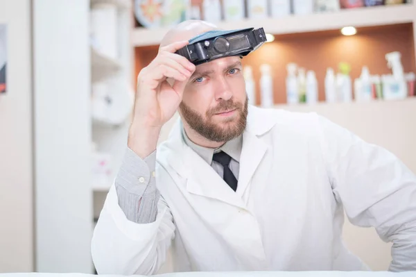 Homem barbudo médico em um casaco médico e com uma alça de cabeça de aumento w luzes sentadas na mesa. Equipamento optometrista. Consultório médico. — Fotografia de Stock