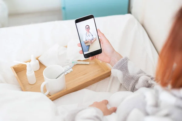Elinde akıllı bir telefonla pijamalarıyla yatakta yatan bir kadın. Bir grip hastası tıbbi video blogu izliyor. Arkadaş canlısı bir doktor telefonla danışmanlık yapıyor.. — Stok fotoğraf