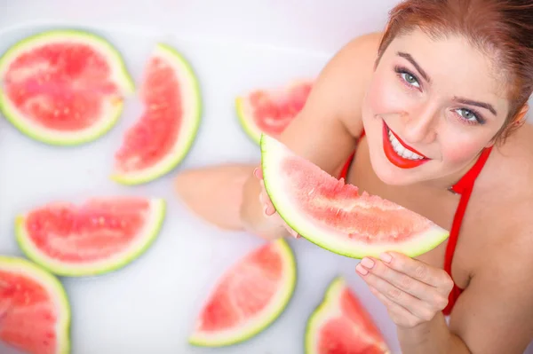 Πορτρέτο μιας γυναίκας με κόκκινο μαγιό που τρώει καρπούζι και χαμογελάει. Redhead κορίτσι με κόκκινο κραγιόν παίρνει ένα μπάνιο με γάλα και φρούτα για να αναζωογονήσει το δέρμα της. Φροντίδα σώματος. — Φωτογραφία Αρχείου