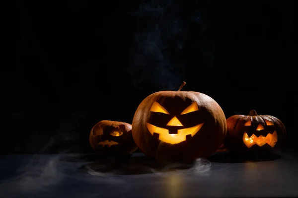 Tři strašidelné halloween dýně s vyřezávaným zářivým úšklebkem na černém pozadí. Ručně vyrobená hlava z lucerny se svíčkou ve tmě mezi mlhou. Trick or treat. Pohlednice. — Stock fotografie