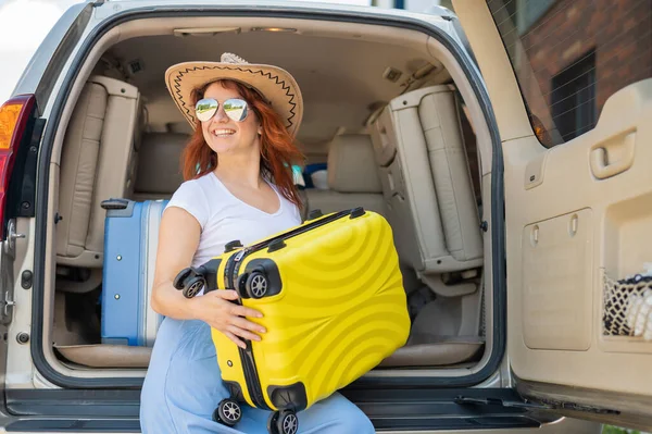 Glückliche rothaarige Frau mit Cowboyhut und Sonnenbrille auf einer Autofahrt durch das Land. Mädchen im Kofferraum mit gelbem Koffer, bereit für den Sommerurlaub. Unabhängiges Reisen. — Stockfoto