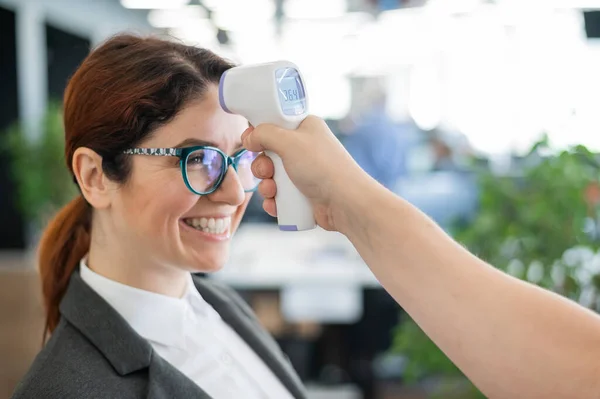 Temperaturmessung mittels elektronischem Infrarotthermometer für alle Büroangestellten. Glückliche Frau im Businessanzug mit elektronischem Thermometer an der Stirn. Coronavirus-Prävention — Stockfoto