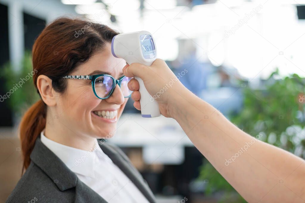 Temperature measurement by infrared electronic digital thermometer to all office workers. Happy woman in a business suit with an electronic thermometer near her forehead. Coronavirus Prevention
