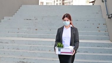 Maskeli mutsuz kadın açık havada merdivenlerin arka planında bir kutu eşyayla cadde boyunca yürüyor. Kadın bir ofis çalışanı kovulmuş. Salgın sırasında ekonomik kriz covid 19.