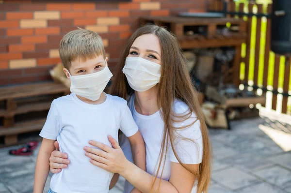 Woman hugs her son in a mask outdoors. Happy little boy with mother are walking in the backyard during isolation. Protection against the spread of coronavirus. Flu outbreak. — Stock Photo, Image