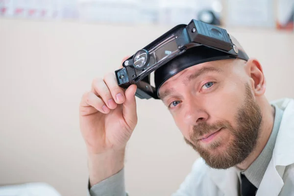 Maschio medico barbuto in un cappotto medico e con una cinghia di ingrandimento w luci seduto sulla scrivania. Attrezzature optometriche. Ufficio medico. — Foto Stock