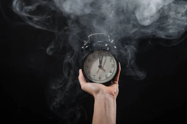 Primer plano de una mano femenina sosteniendo un reloj sobre un fondo negro en humo. Reloj despertador a medianoche en una niebla mística . —  Fotos de Stock