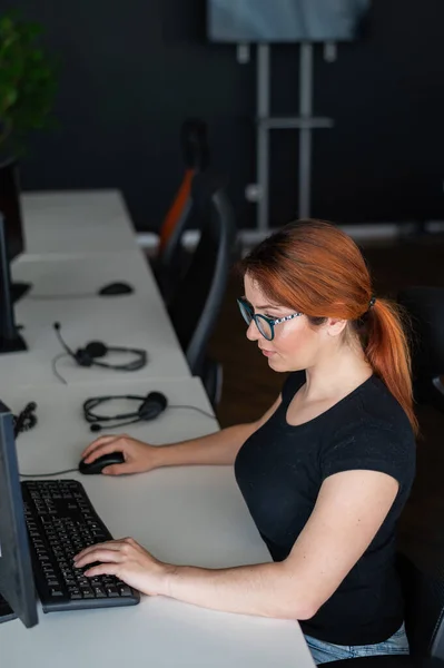 안경을 쓰고 평상복 차림의 빨간 머리 여자는 시간외 근무를 한다. 초점을 맞춘 소녀 혼자 빈 사무실에 있는 컴퓨터에서 일 한다. 데드 라인. IT 전문가가 주말에 열심 히 일 한다. — 스톡 사진
