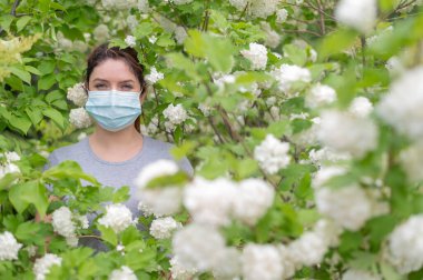 Sağlık maskeli, mutsuz, kızıl saçlı bir kadın parkta çiçek açan elma ağacının yanında duruyor. Mevsimsel alerjik rhinitis. Kızın polene alerjisi var..