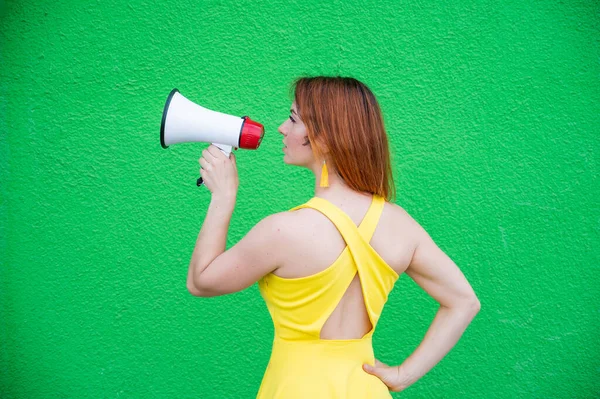 Razhevolosaya belle femme en robe jaune debout derrière et criant dans un mégaphone. Fille fait une annonce sur le haut-parleur sur fond de mur vert. Vue arrière. — Photo