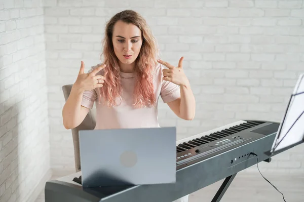 Uma mulher amigável toca piano eletrônico e conduz um vídeo blog em seu laptop. Fica em casa. Professor de instrumentos musicais. Música de aprendizagem à distância em quarentena. — Fotografia de Stock