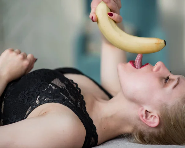 A loira em roupa interior de renda preta eroticamente come uma banana. Mulher atraente com lábios vermelhos sensuais suga sexualmente e lambe uma banana. Prazer oral. — Fotografia de Stock